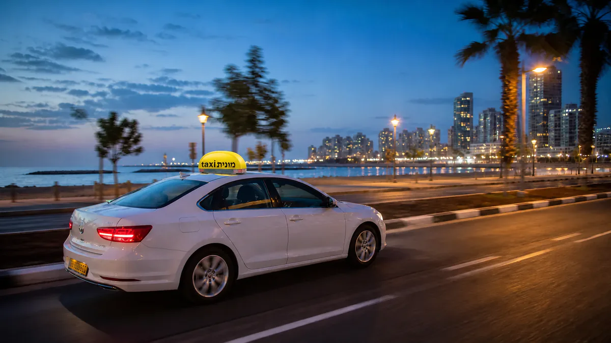 מונית לבנה עם כובע צהוב ועליו הכיתוב מונית taxi לאחר נסיעה מנתב״ג לתל אביב־יפו